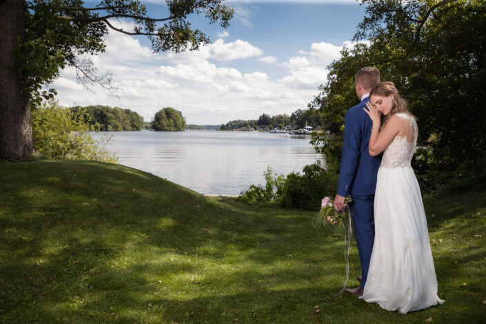 Kreative Hochzeitsbilder vom Hochzeitsfotograf Berlin: Braut umarmt von hinten den Bräutigam, im Hintergrund. der Lehnitzsee