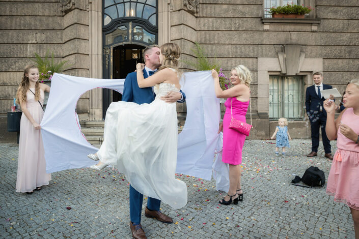 Bild vom Hochzeitsfotograf Berlin aus der Hochzeitsreportage -hier abgebildet: Bräutigam trägt Braut durch das zerschnittene Bettlaken vor dem Standesamt Potsdam